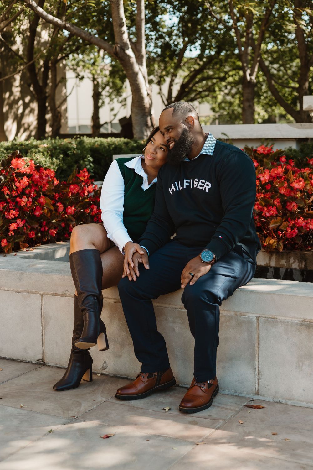 A couple sits together on a concrete ledge with red flowers blooming in the background, showing their matching brown boots.