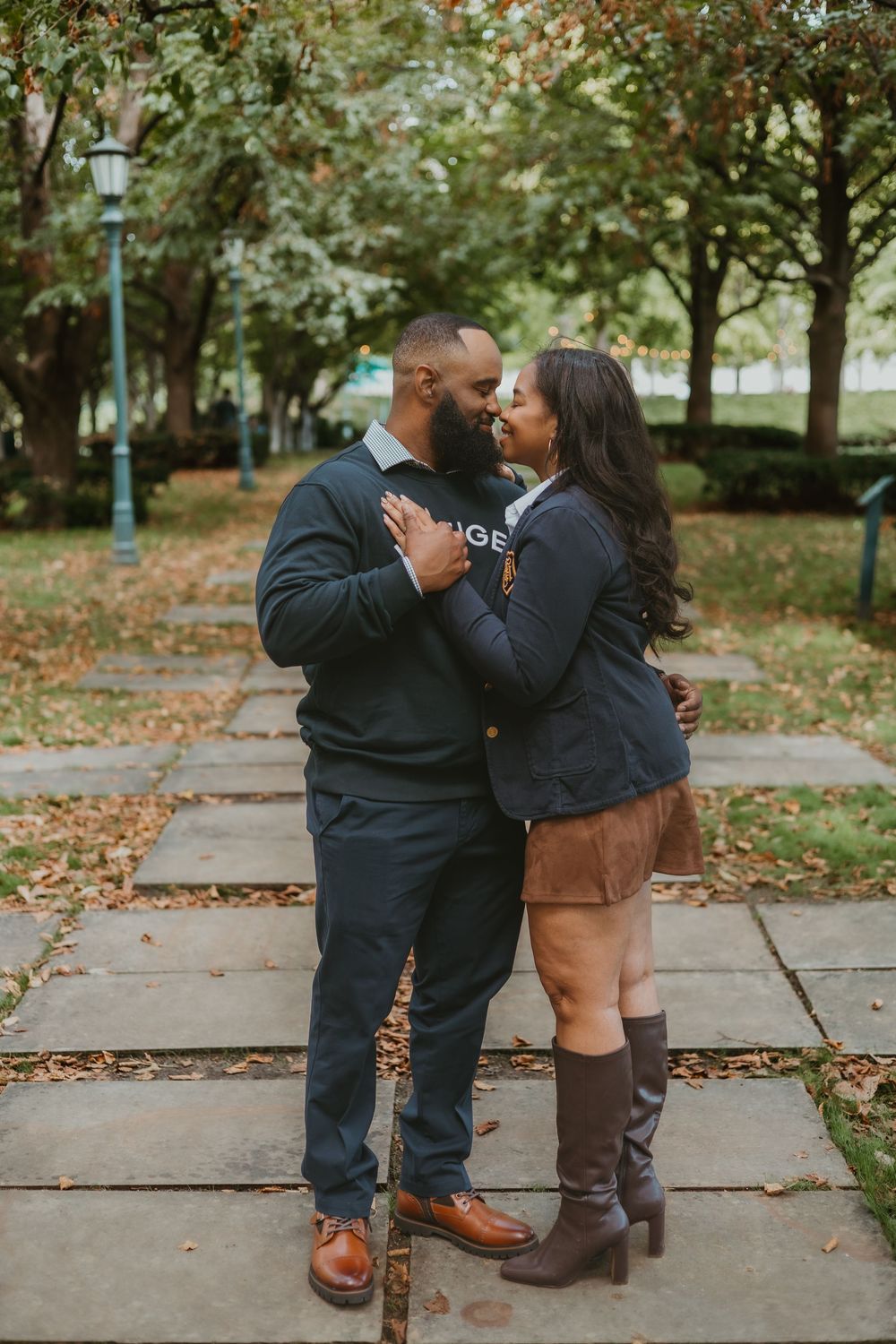 A couple shares an intimate moment on a path lined with autumn trees and fallen leaves.