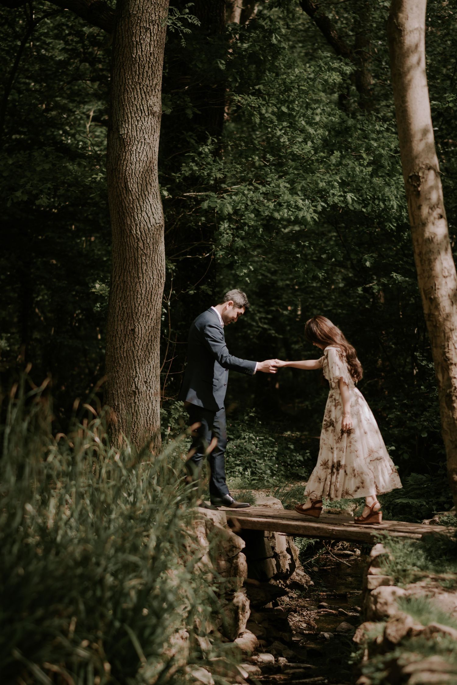 A couple holds hands while balancing on a rustic wooden bridge in a lush forest.