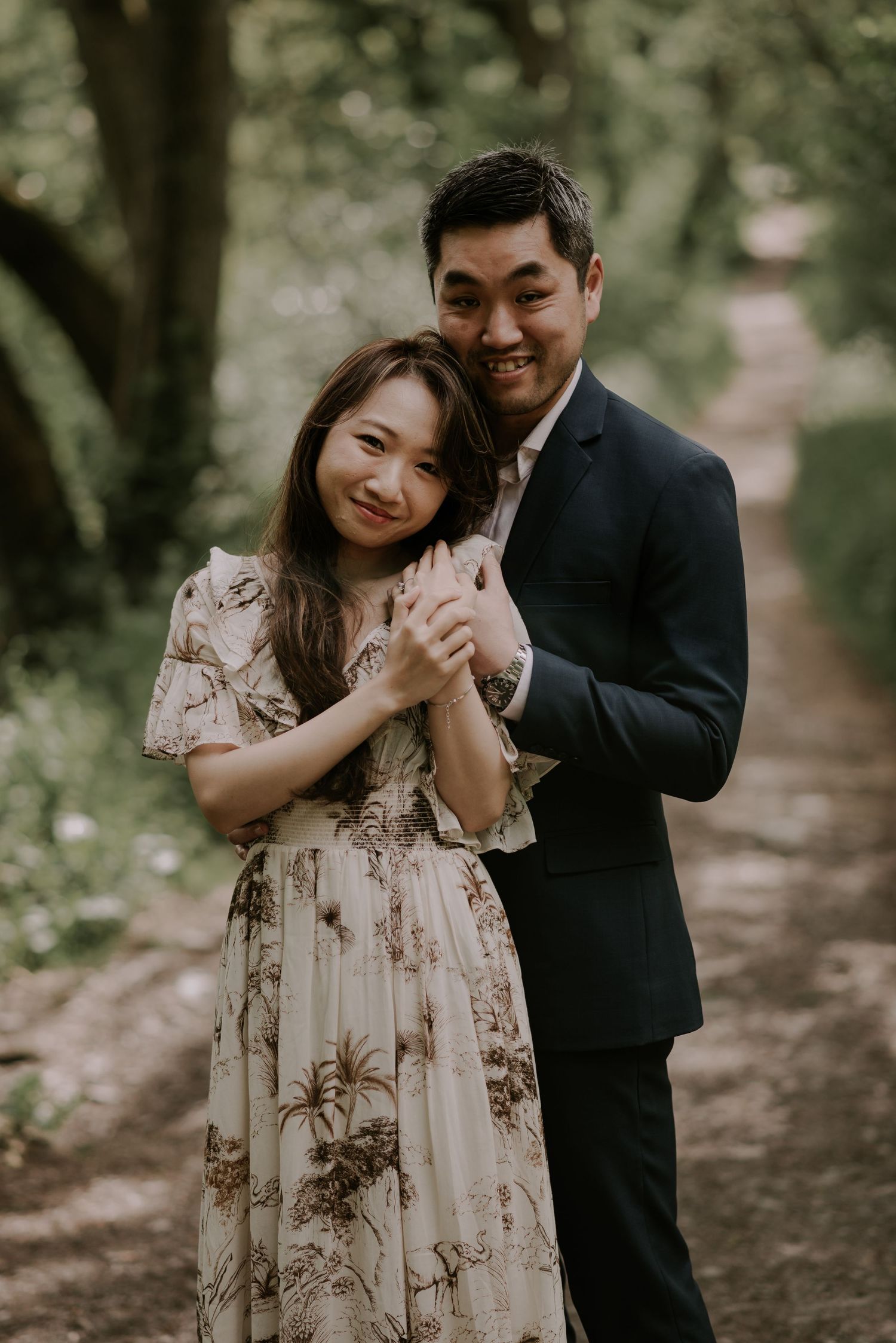 A couple in formal attire embraces on a woodland path during a romantic photoshoot.