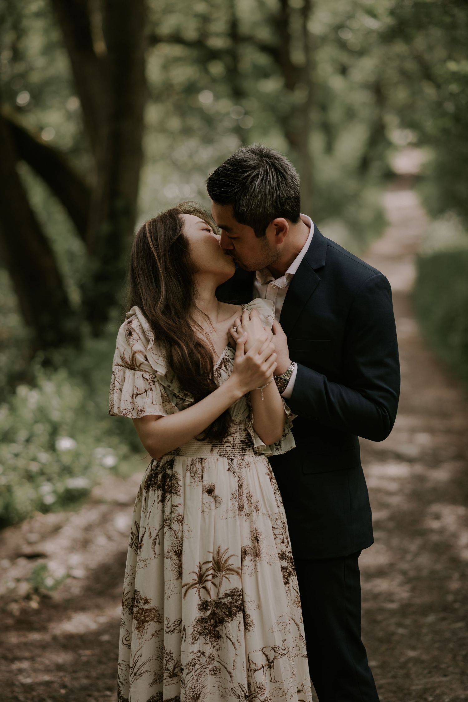 Romantic couple embraces in a wooded path while wearing dressy attire and a floral dress.
