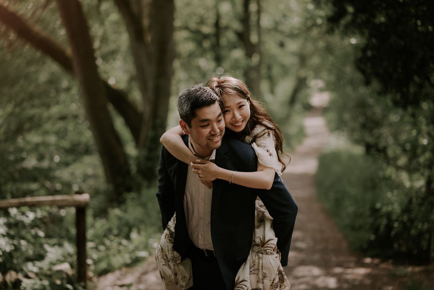 A romantic couple share a playful piggyback ride moment along a tree-lined path.