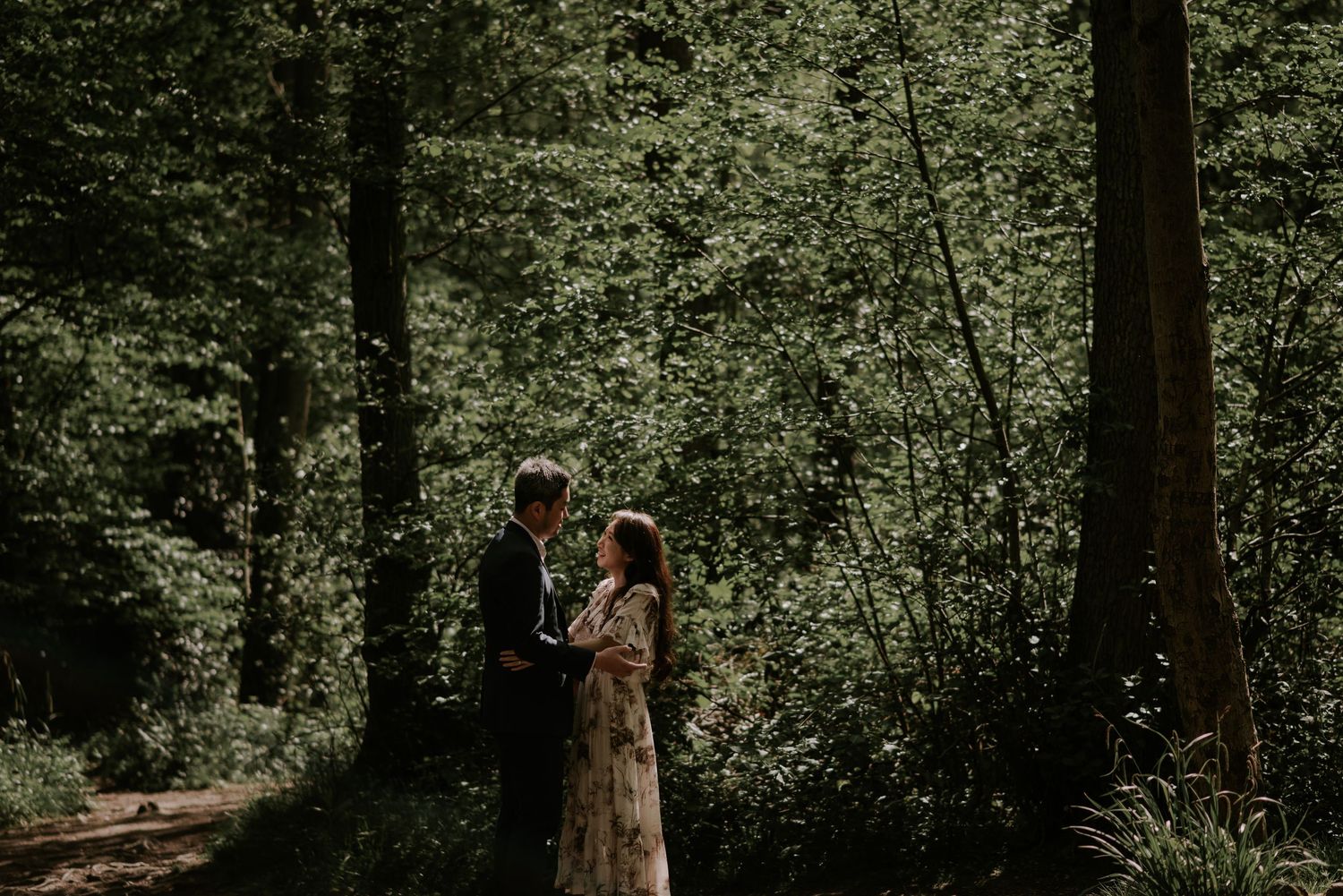 A couple stands together in a sunlit forest clearing during their engagement session.