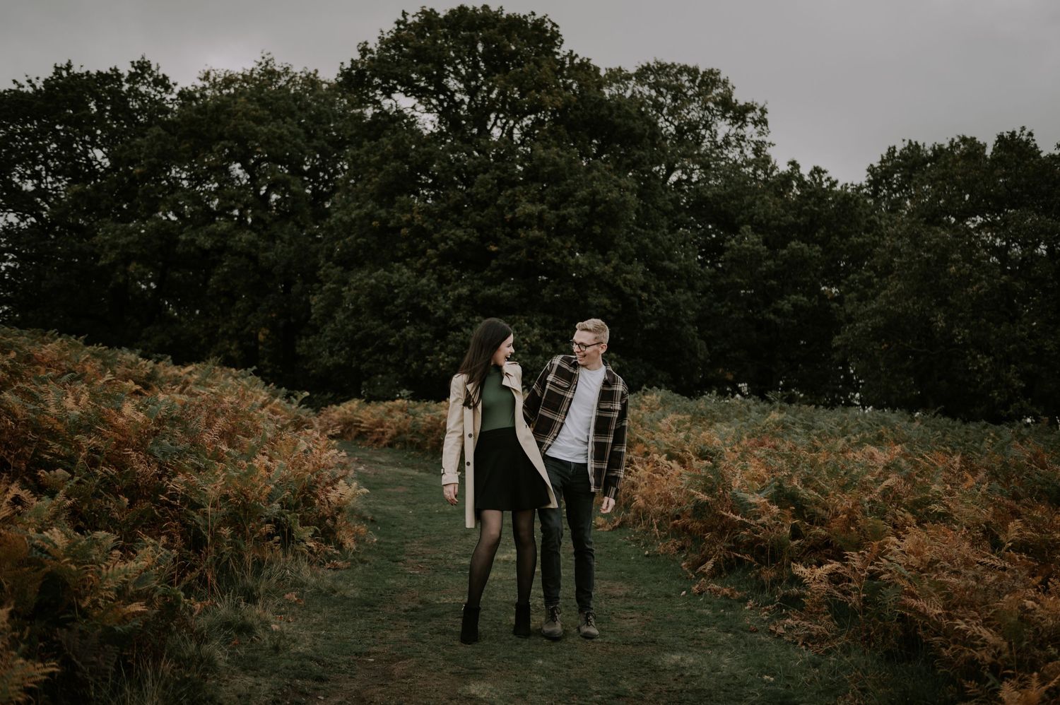 Two people stand together on a narrow path surrounded by autumn ferns and dark woods in moody overcast lighting.