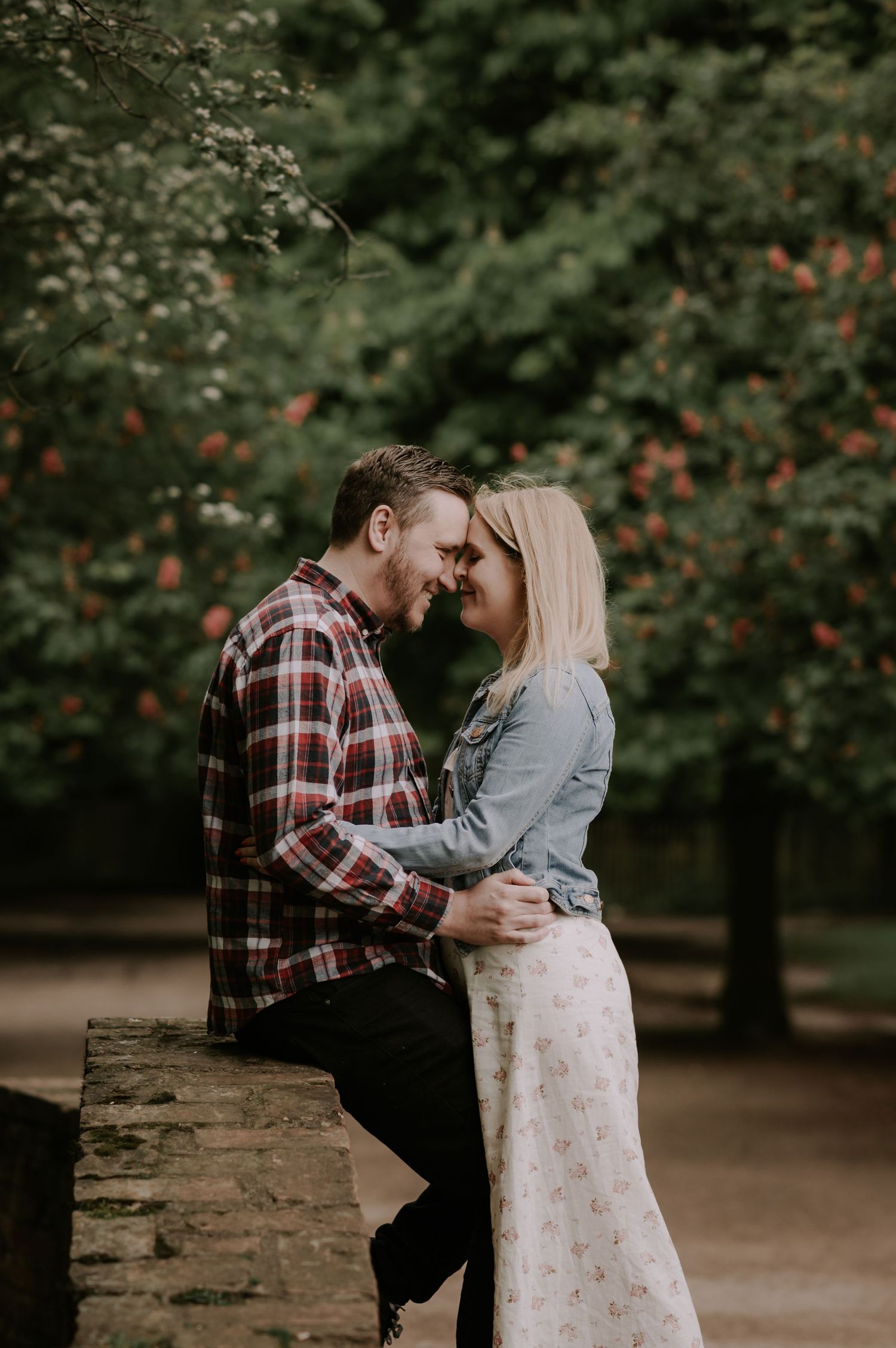 A romantic couple shares an intimate moment on a garden wall surrounded by lush greenery and blooming flowers.