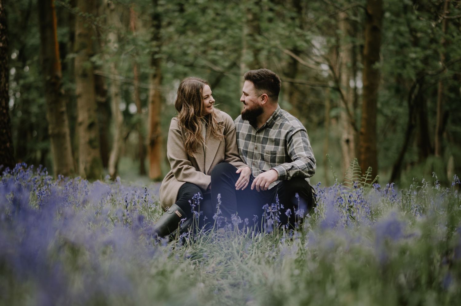 A couple sits together in a blooming field of purple wildflowers surrounded by a tranquil forest setting.