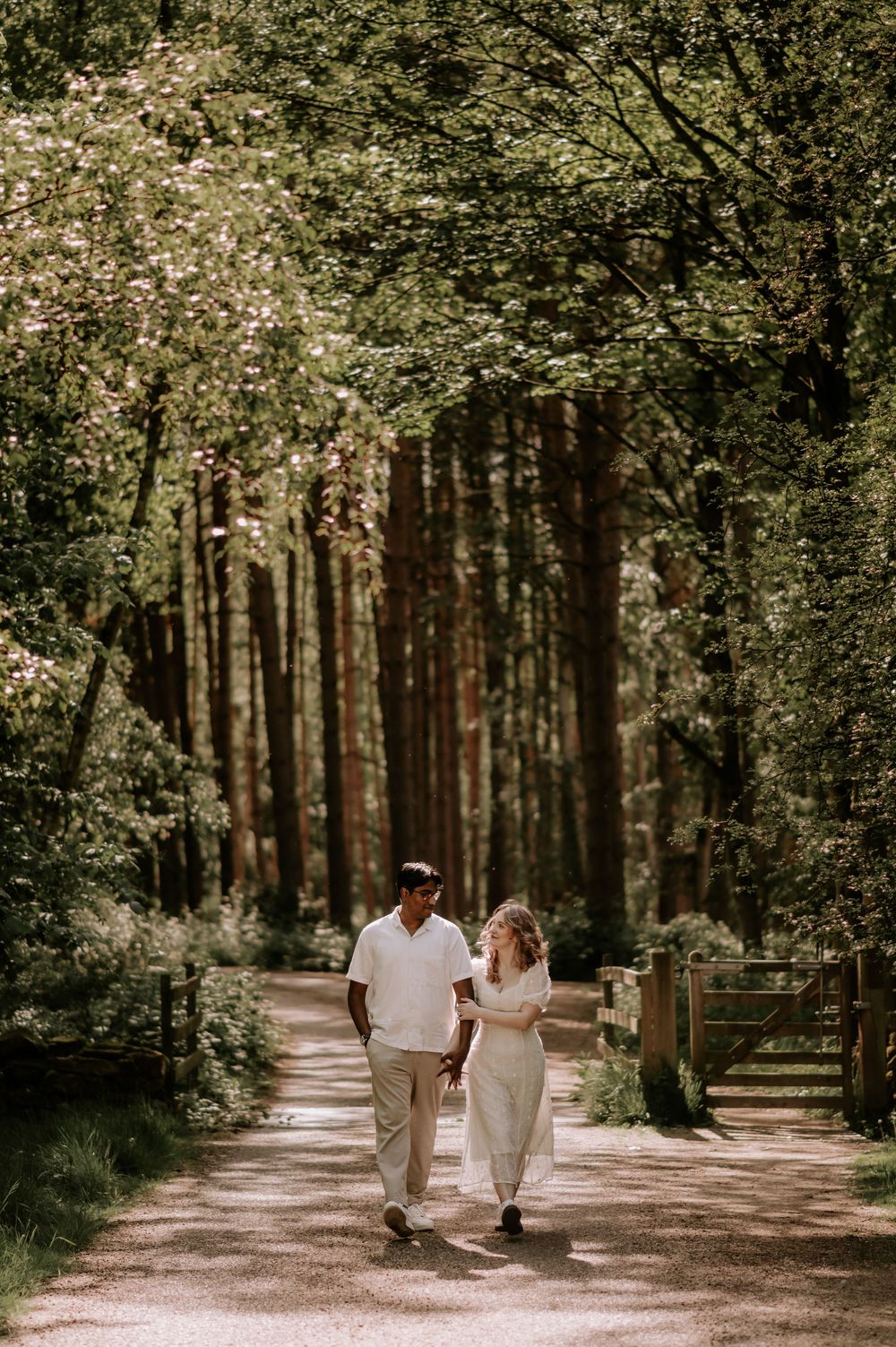 Couple walking hand in hand along a sunlit forest path surrounded by tall trees and lush greenery.