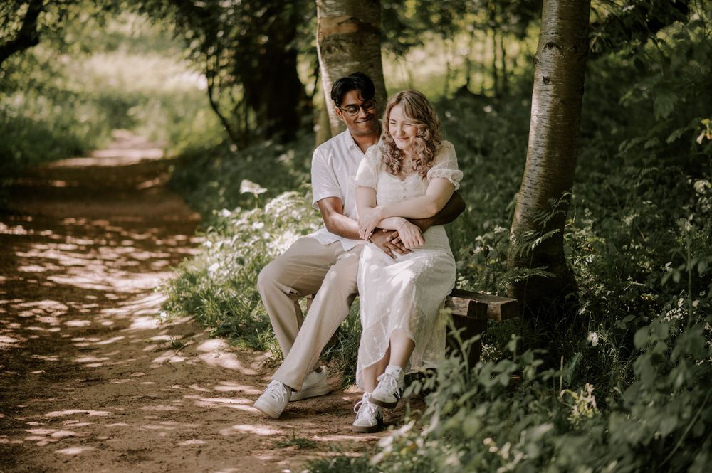 A couple in wedding attire sits on a bench along a sunlit forest path surrounded by lush greenery.