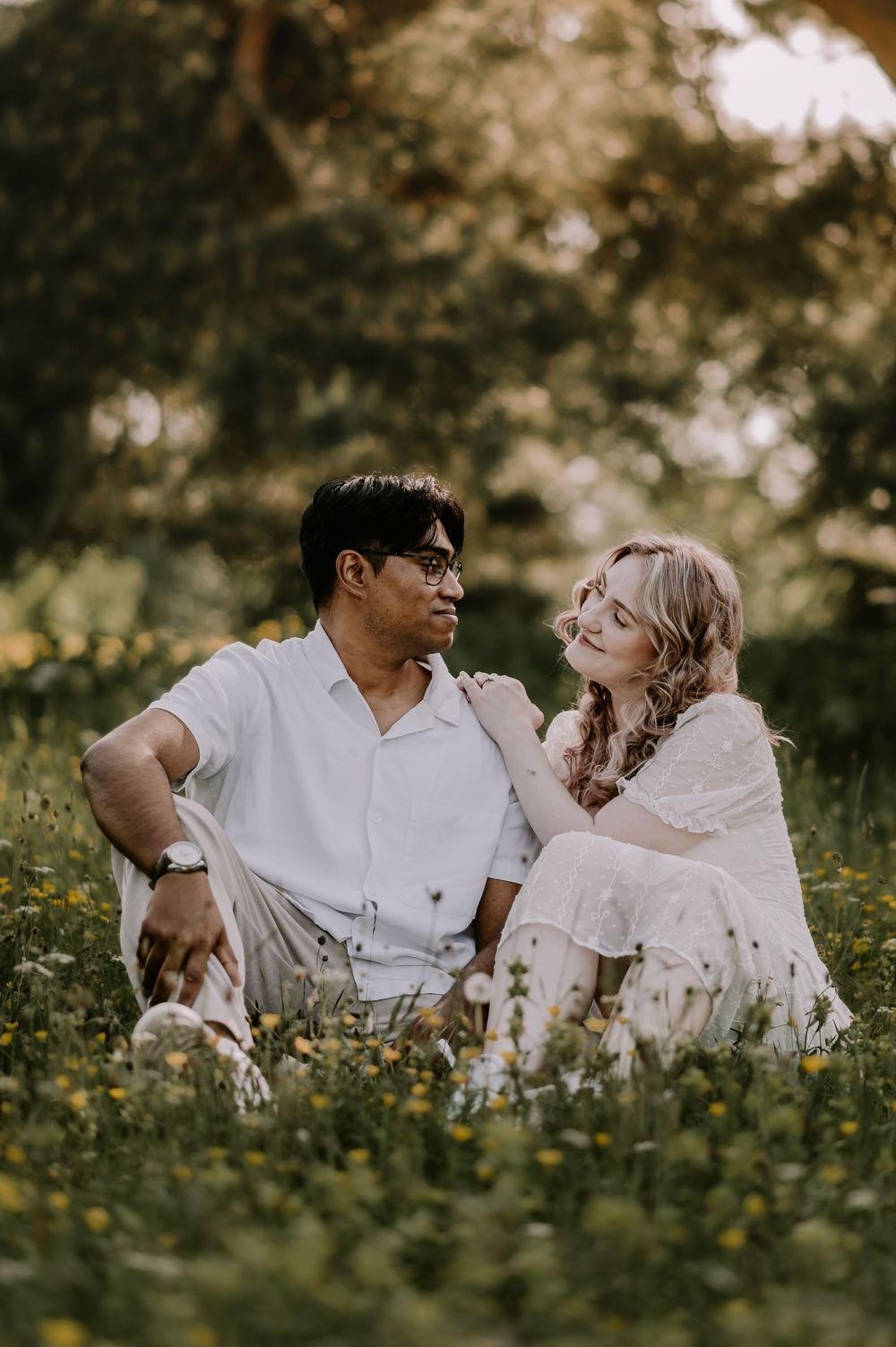 Couple sitting together in a flower meadow, wearing white outfits, sharing a tender moment surrounded by golden sunlight.