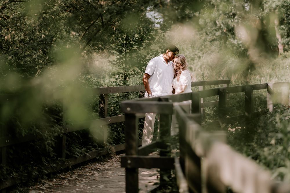 Couple sharing a romantic moment on a wooden bridge surrounded by lush green trees in a serene outdoor setting.