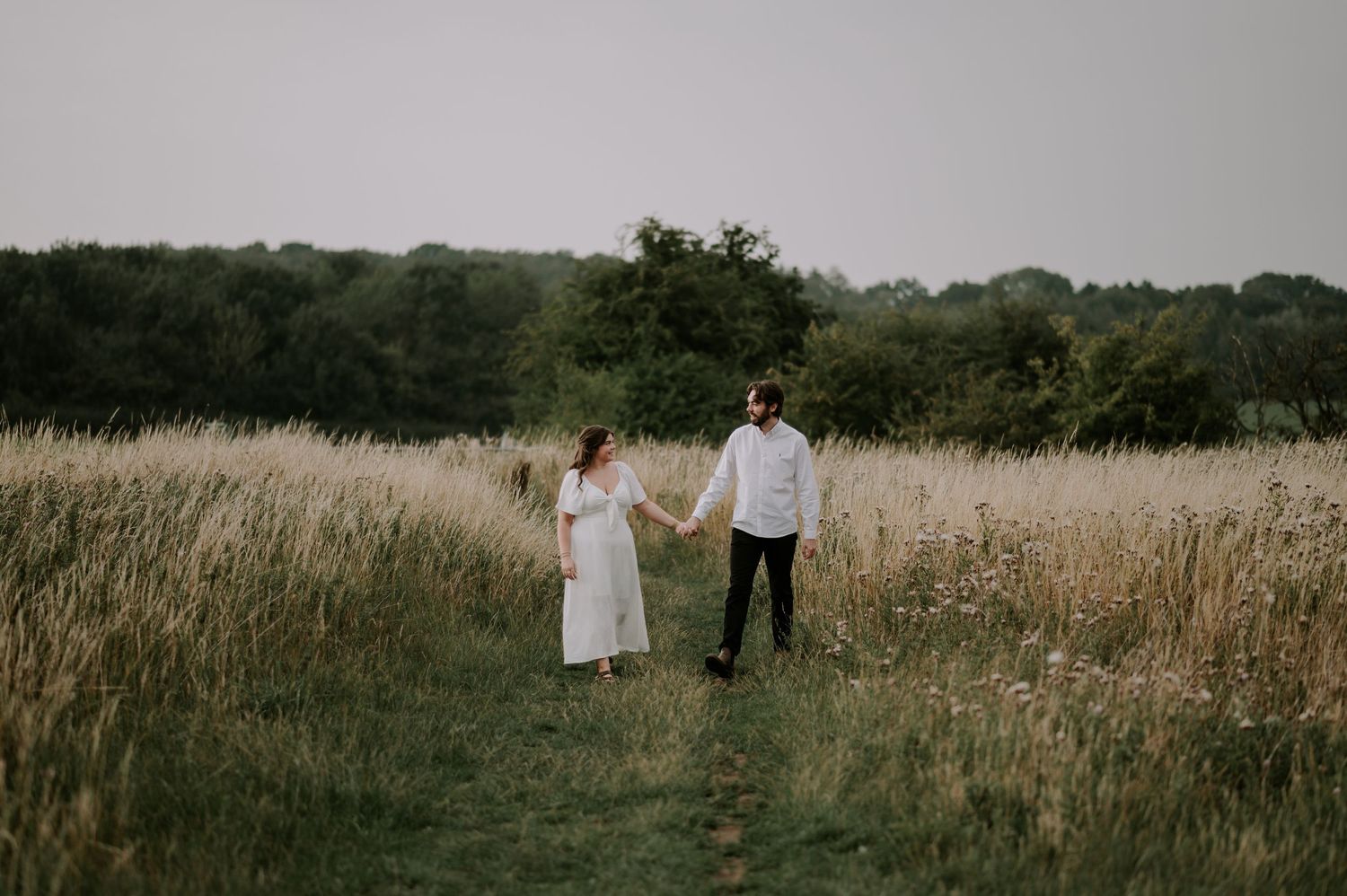 A couple holds hands while standing in a golden meadow at dusk.