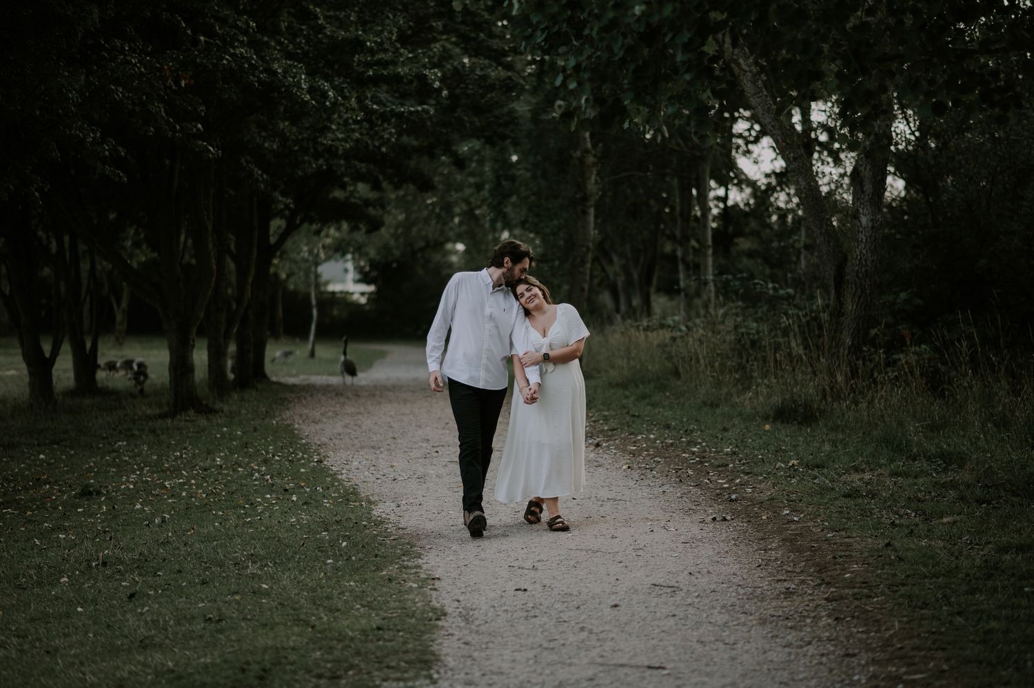 A couple walking together on a tree-lined path during a moody evening photoshoot.