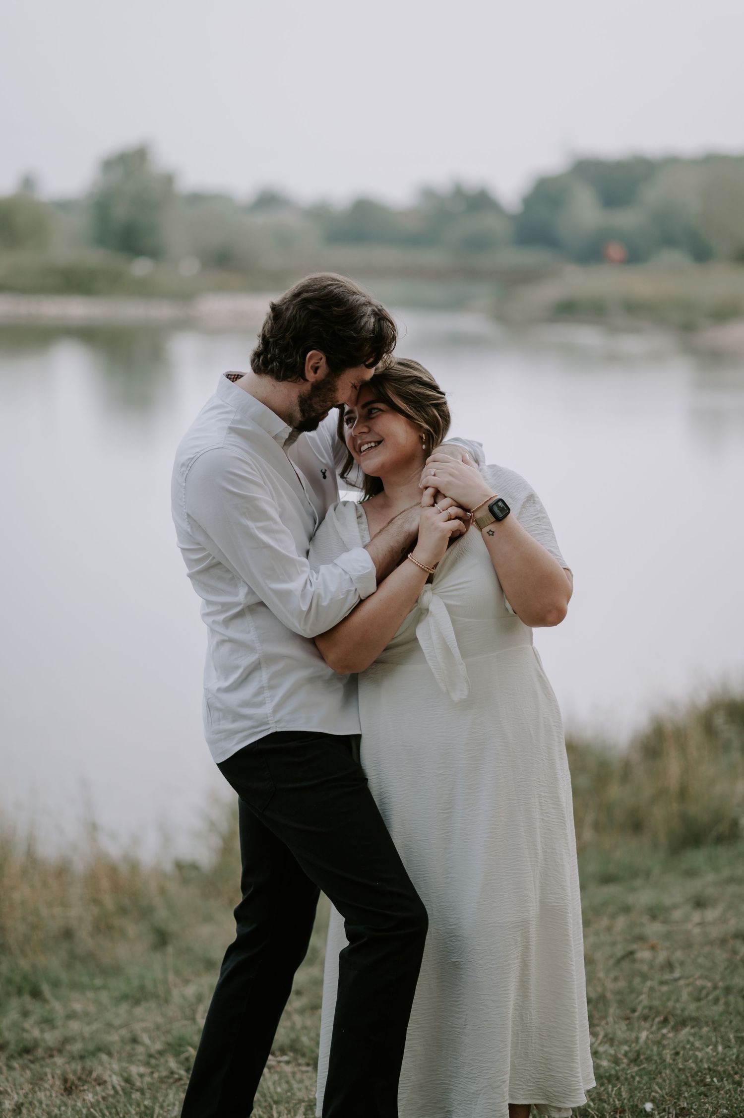 Couple shares intimate moment by lakeside in casual attire during overcast day.