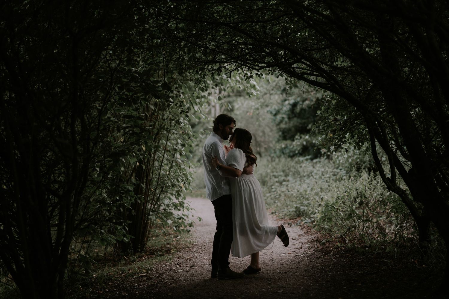 A romantic moment between two figures beneath a natural archway of trees and foliage.