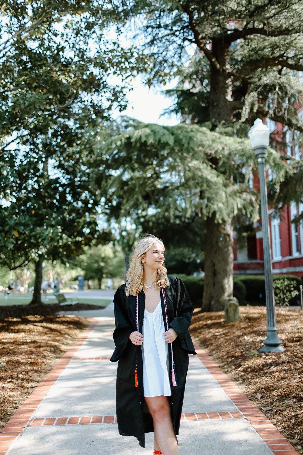 Person in black blazer walks along brick pathway under tree canopy on sunny day.