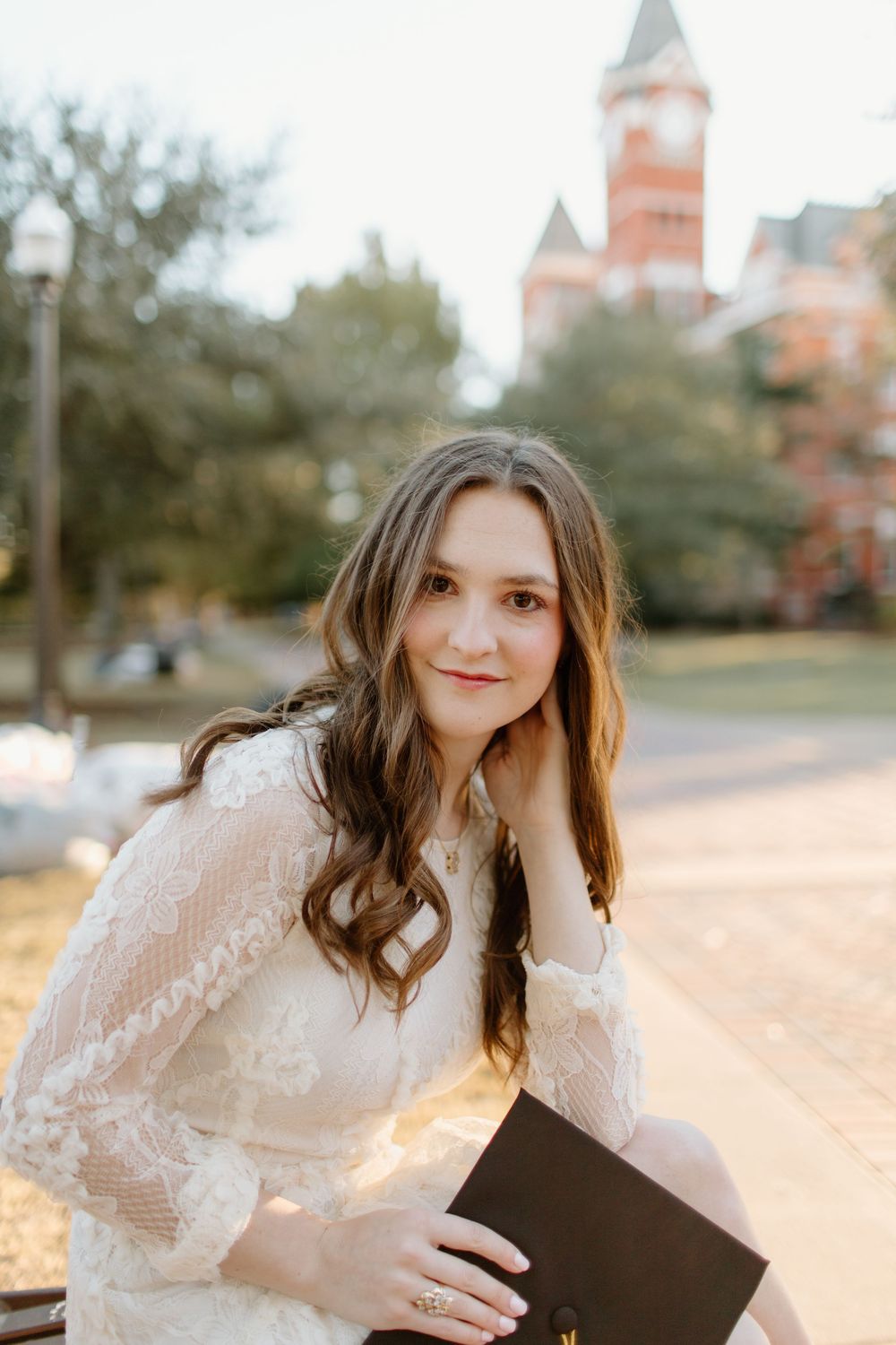 Graduate in white lace dress poses with diploma on campus grounds.