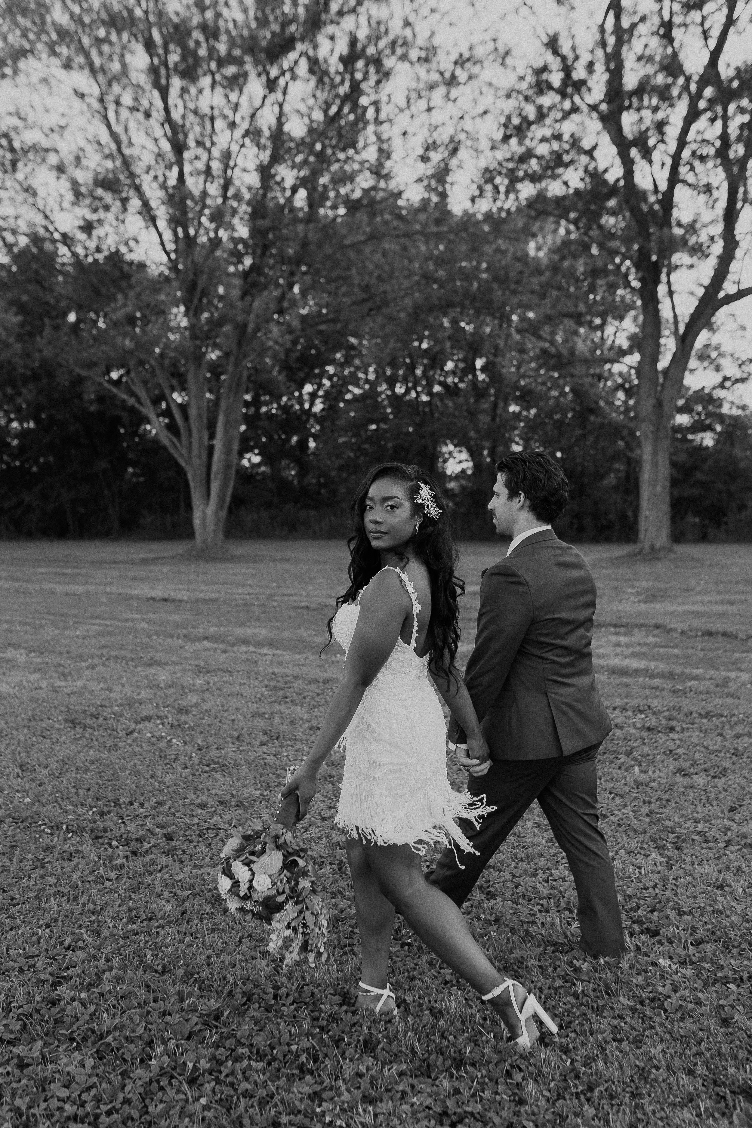 Black and white photo of couple walking playfully with bouquet in outdoor garden setting.