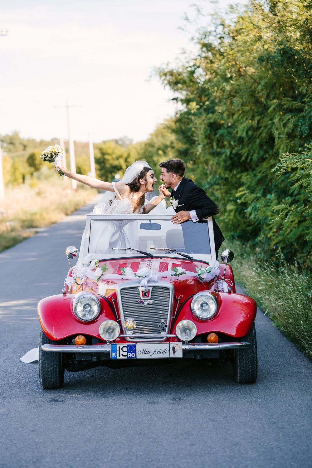 Wedding couple poses playfully in vintage red convertible car on rural road.