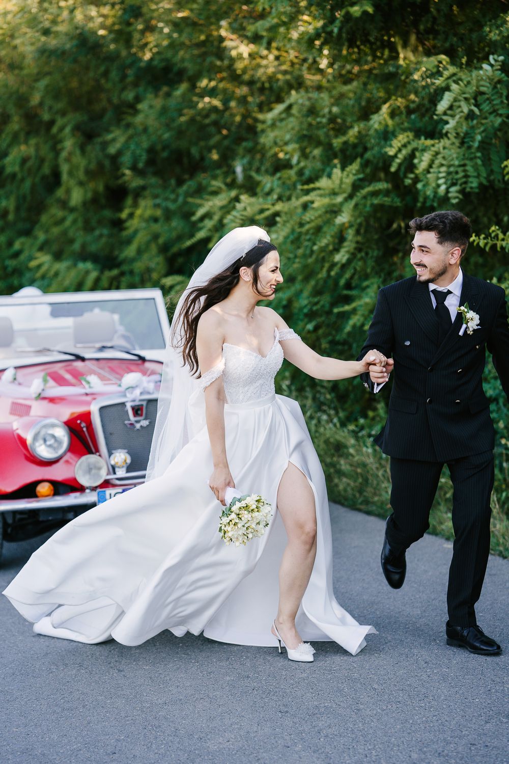 Bride in flowing white dress dances with groom on asphalt road near vintage car.
