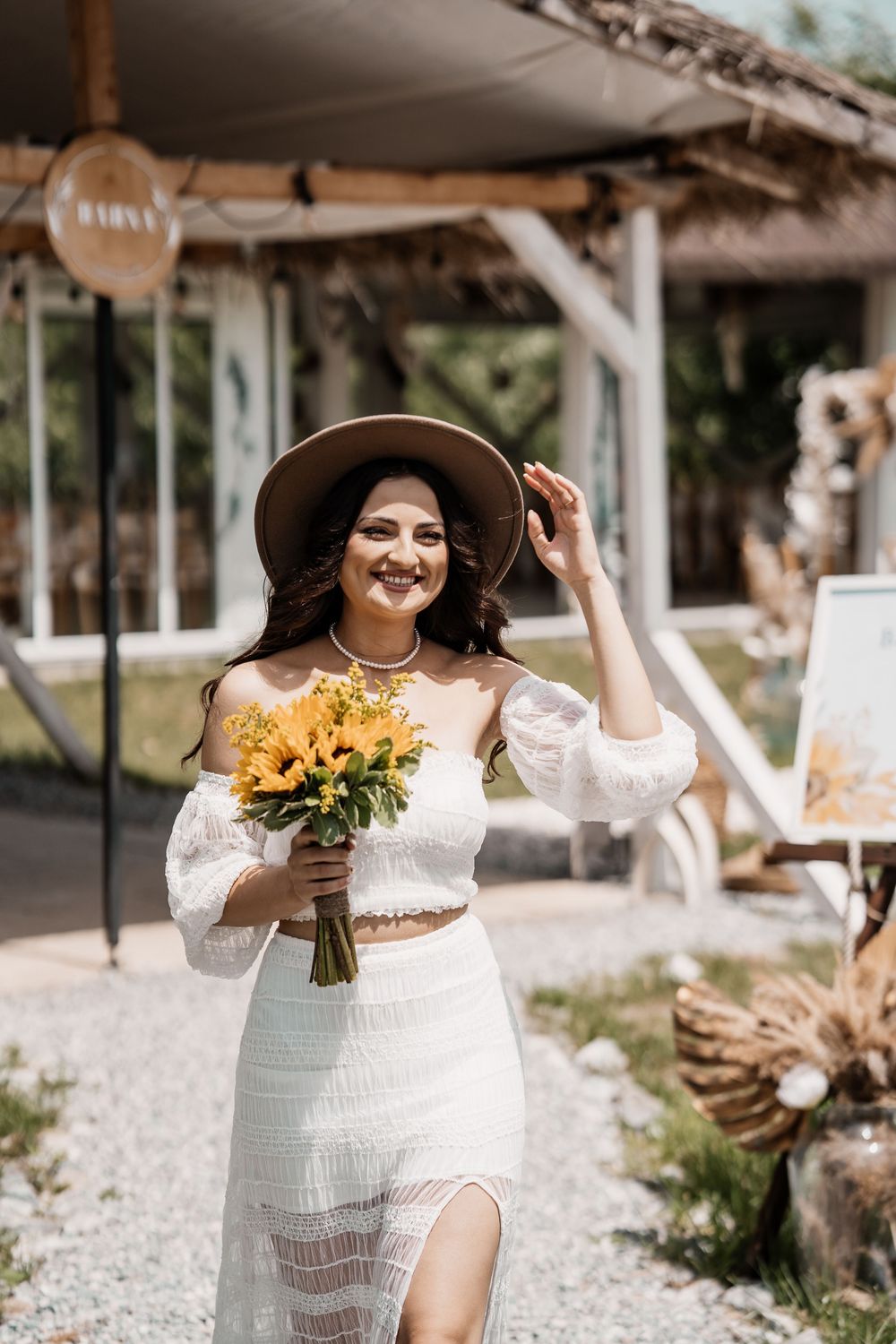 A boho bride in a white lace dress poses with a sunflower bouquet against a rustic backdrop.