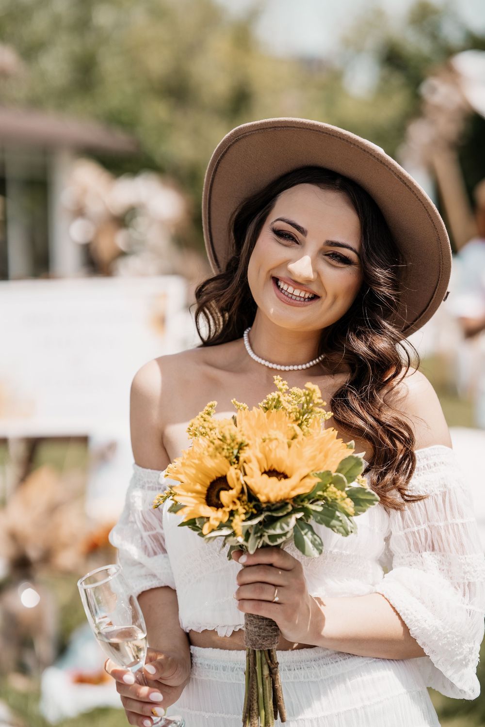 A woman in a white dress and wide-brimmed hat holds a bright sunflower bouquet while smiling.