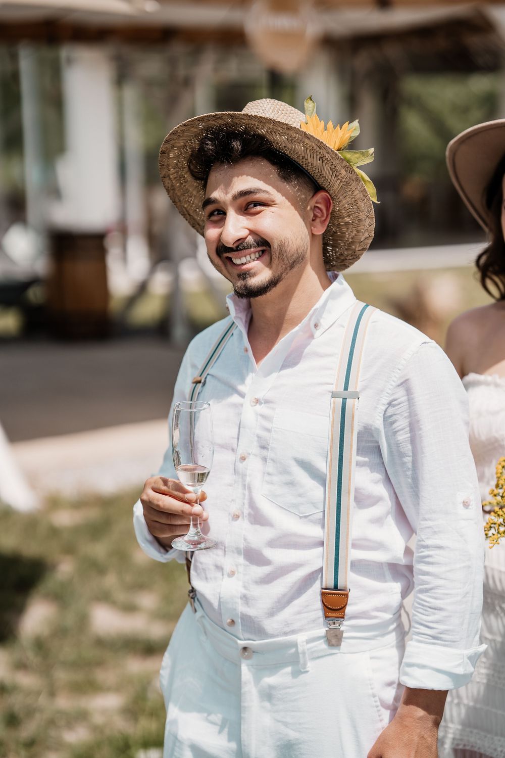 A person in a white shirt and suspenders smiles while wearing a straw hat at an outdoor event.