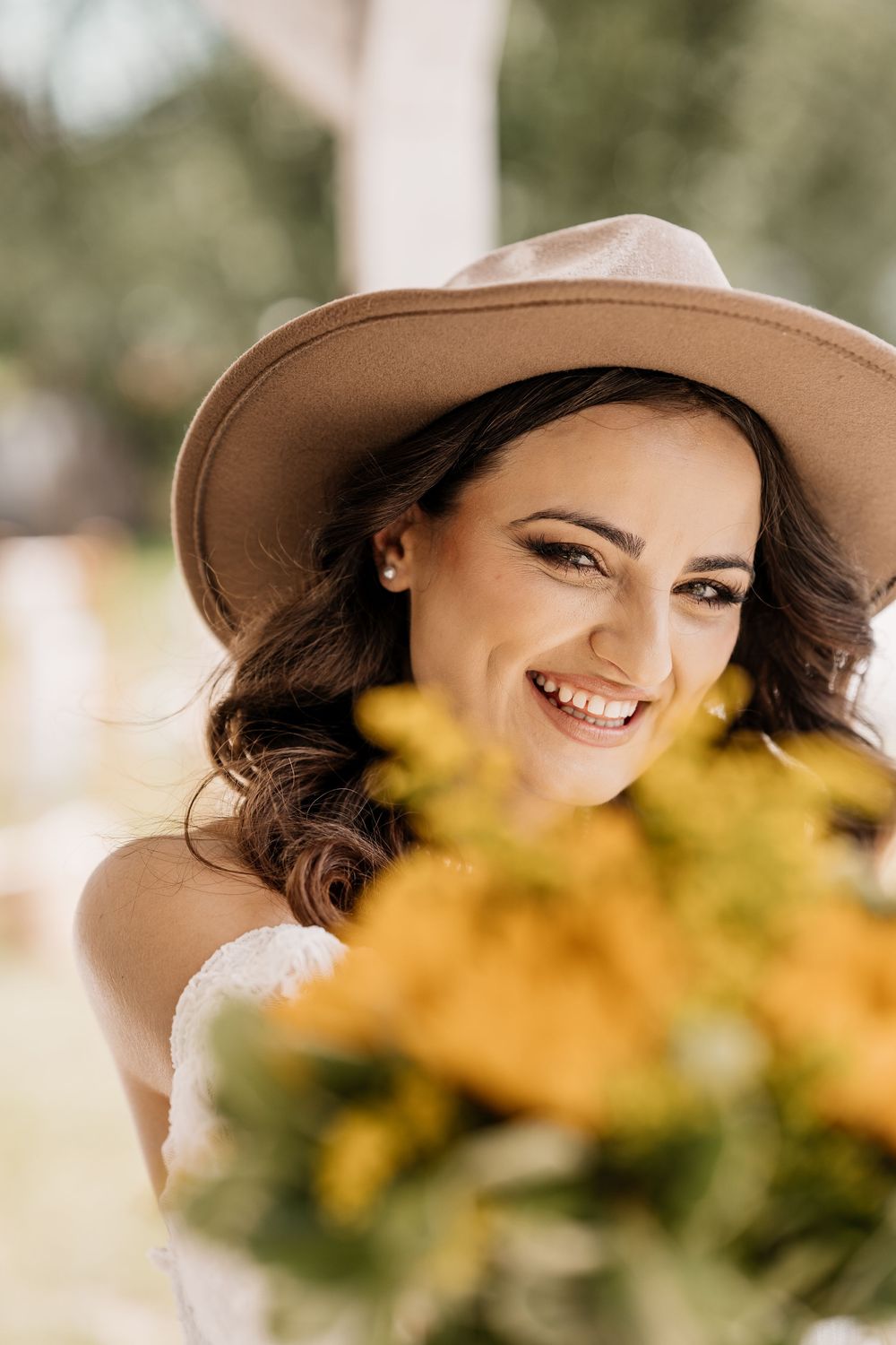 A woman in a brown hat smiles radiantly while posing with yellow flowers in the background.