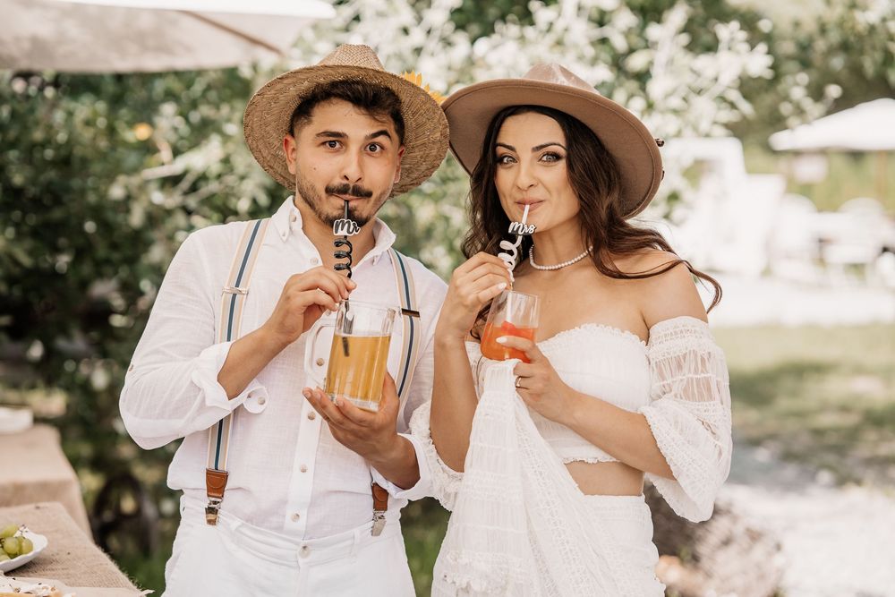 Couple in white attire drinking colorful cocktails at outdoor wedding celebration.