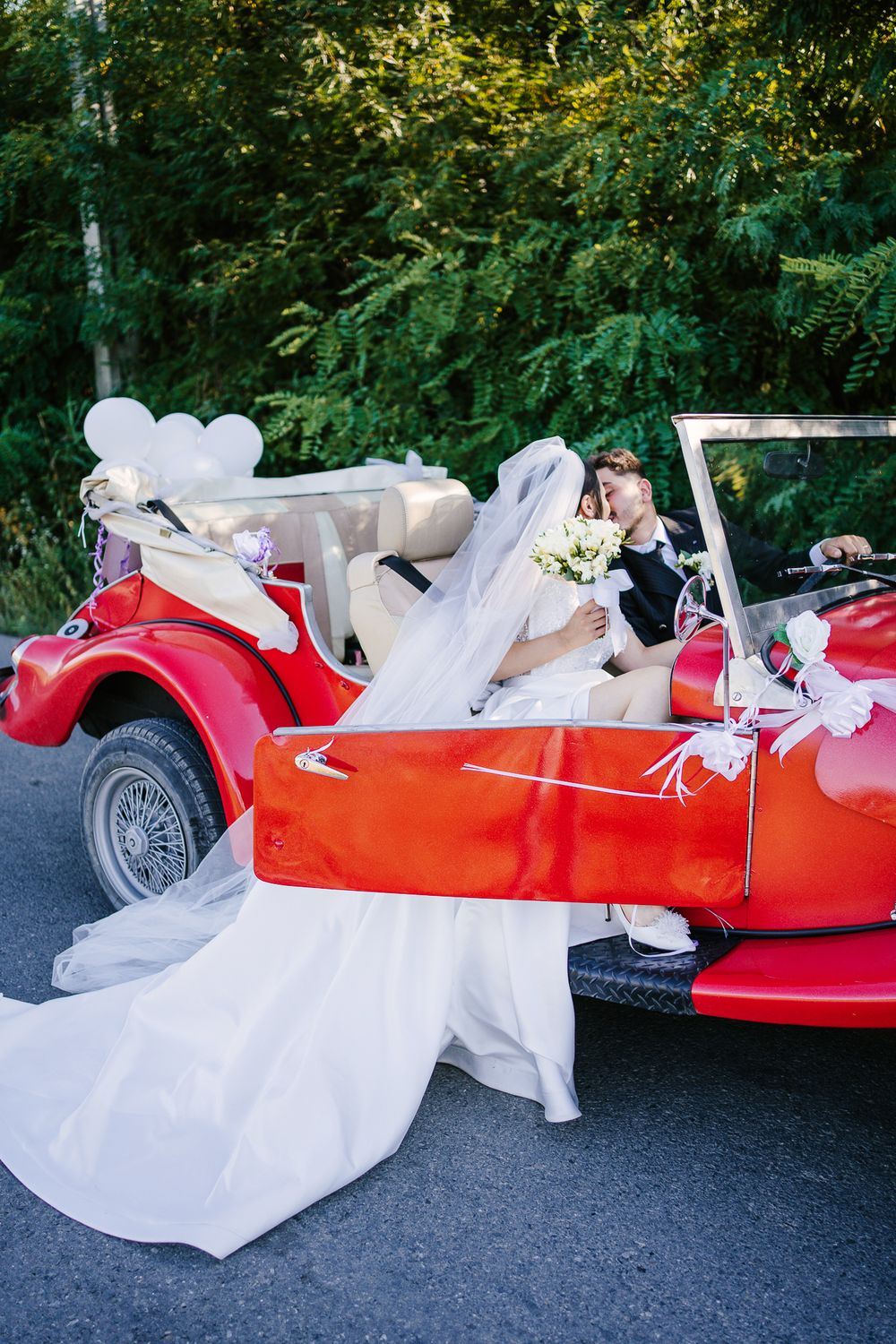 Wedding couple in red classic car with flowing white dress creating dramatic movement.