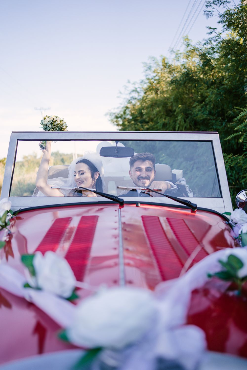 Newlyweds sit in red vintage car decorated with white flowers and ribbons.