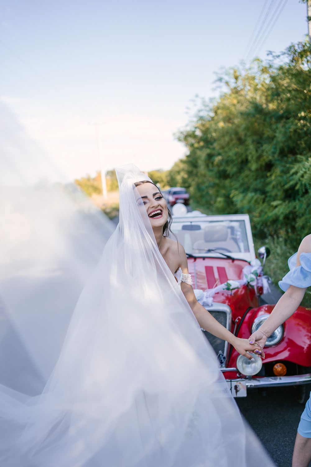 A bride laughs joyfully in her flowing white dress while leaning back during an outdoor celebration.