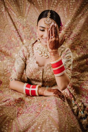 Picture of the bride with her one hand covering her face and the background consists of lehenga of the bride.