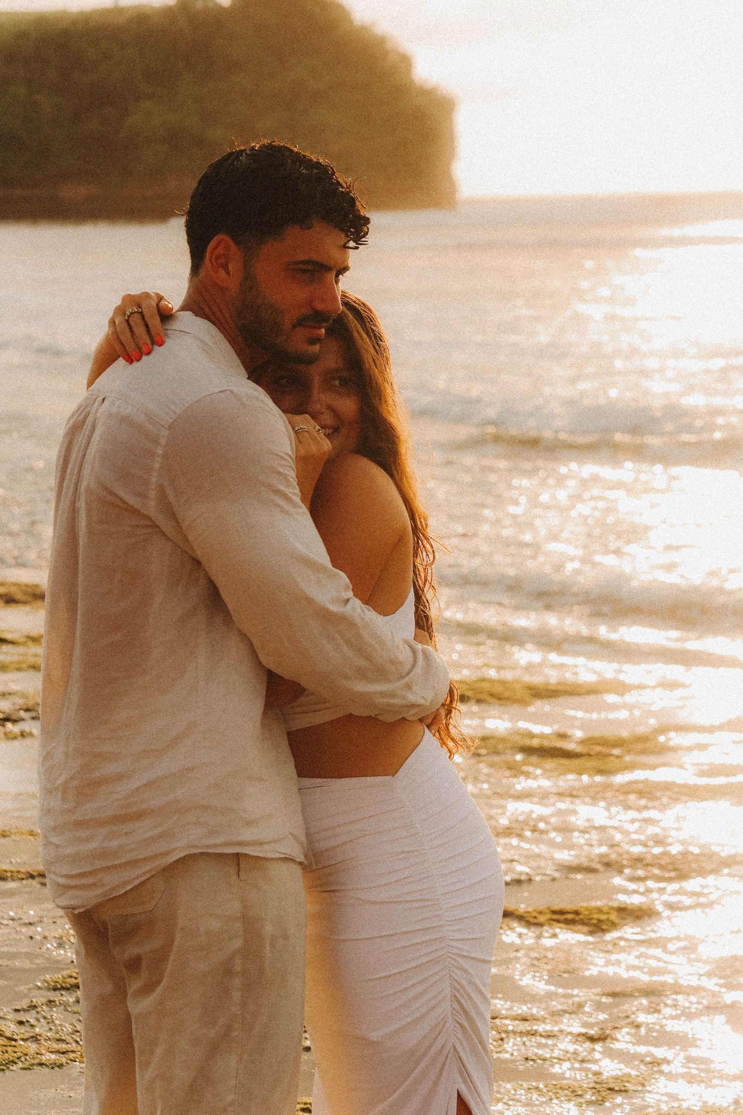Romantic couple hugging on beach at golden sunset, woman in white dress smiling over mans shoulder.