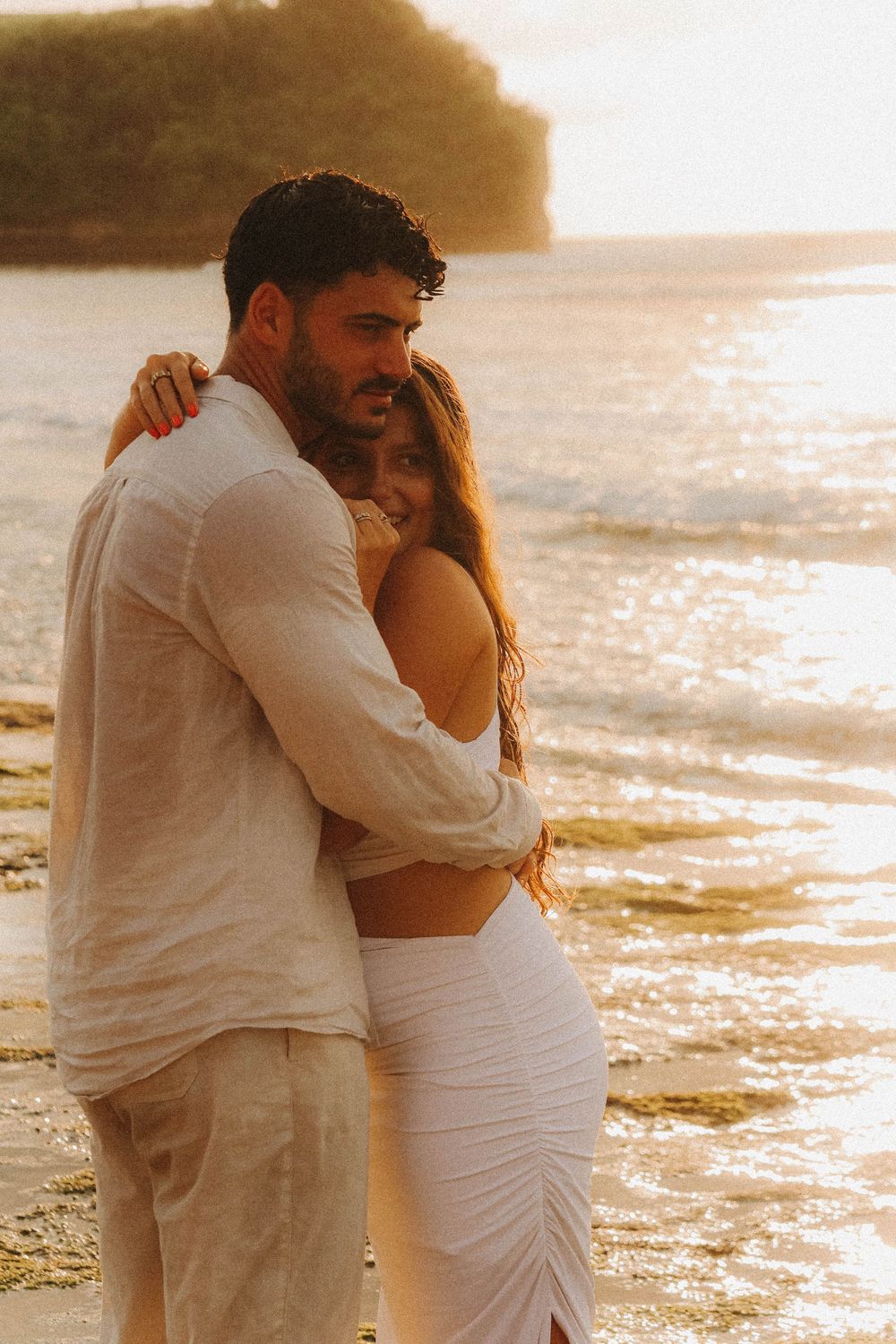 Romantic couple hugging on beach at golden sunset, woman in white dress smiling over mans shoulder.