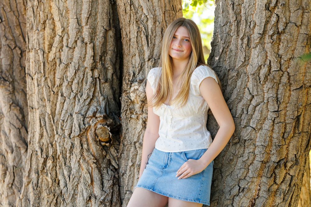 Young woman in white top and denim skirt leaning against large tree trunk outdoors.