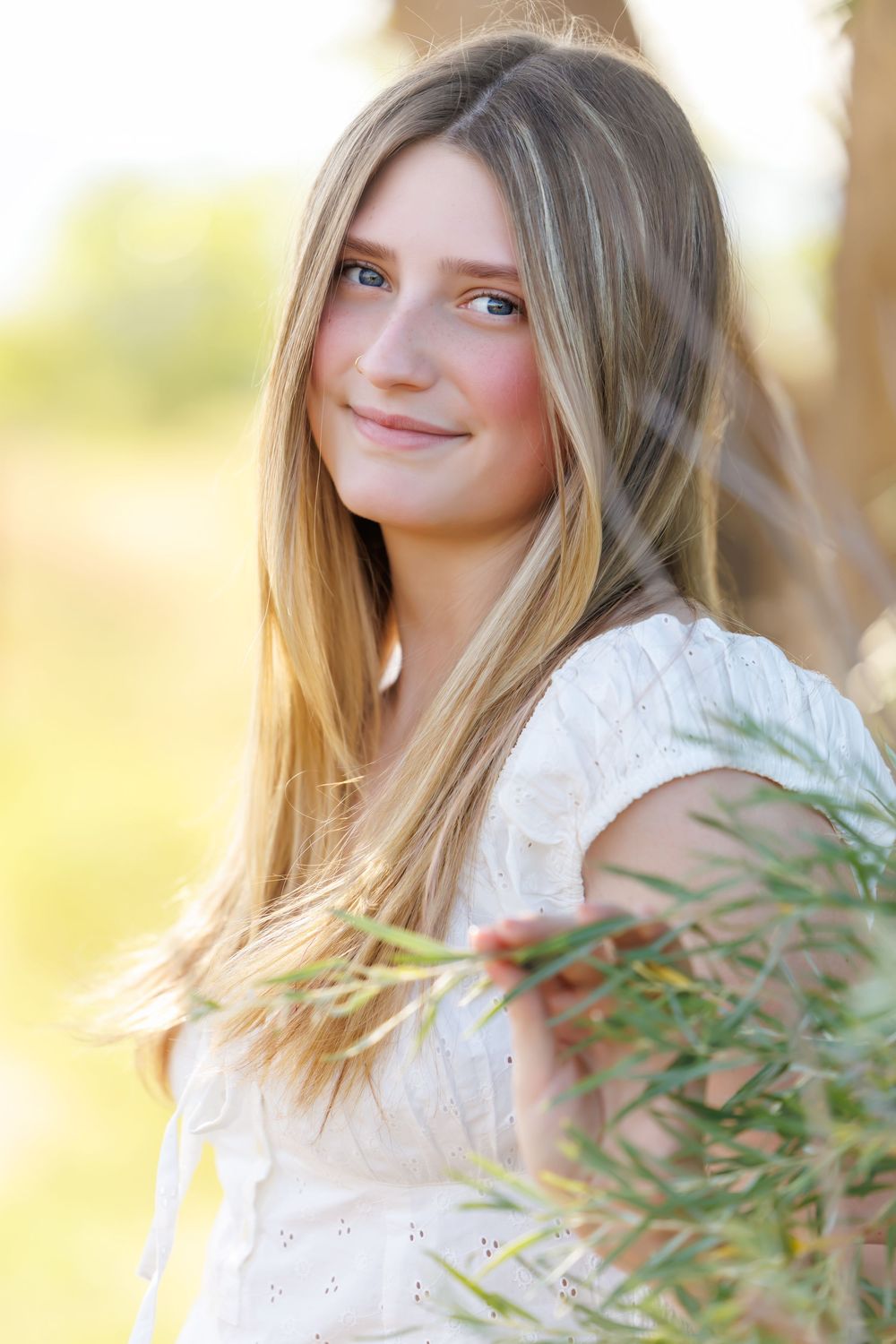 Young blonde woman in white dress holding green branches outdoors in golden sunlight.