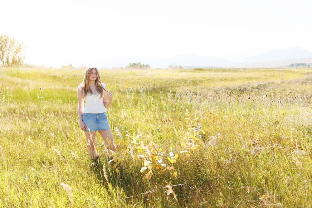 Young woman in white top and denim shorts standing in a sunny golden meadow with mountains in the background.