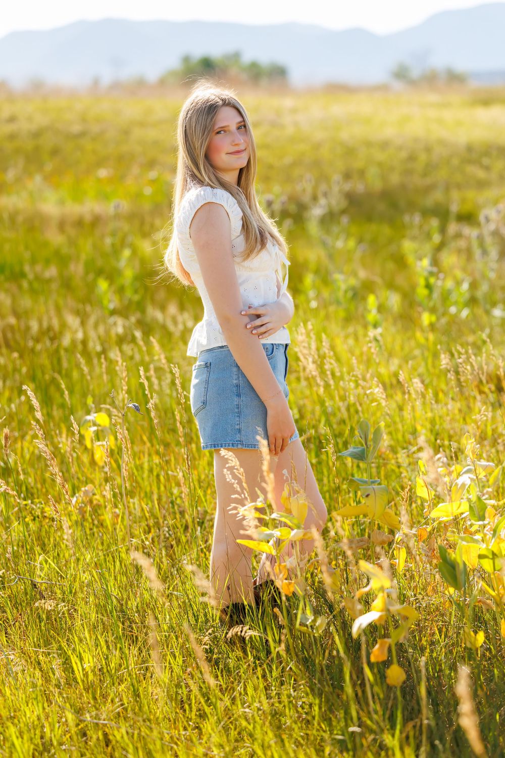 Young blonde woman in white top and denim shorts standing in sunny green meadow with mountains in background.