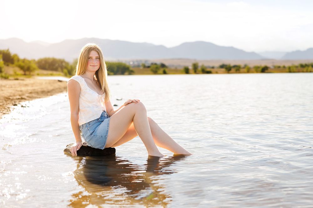 Young woman in white top and denim skirt sitting by a lake shore with mountains in the background.
