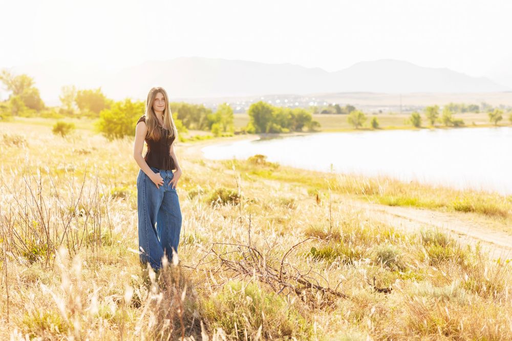Young woman in blue jeans and black top standing in golden field with mountain lake backdrop in sunlight.