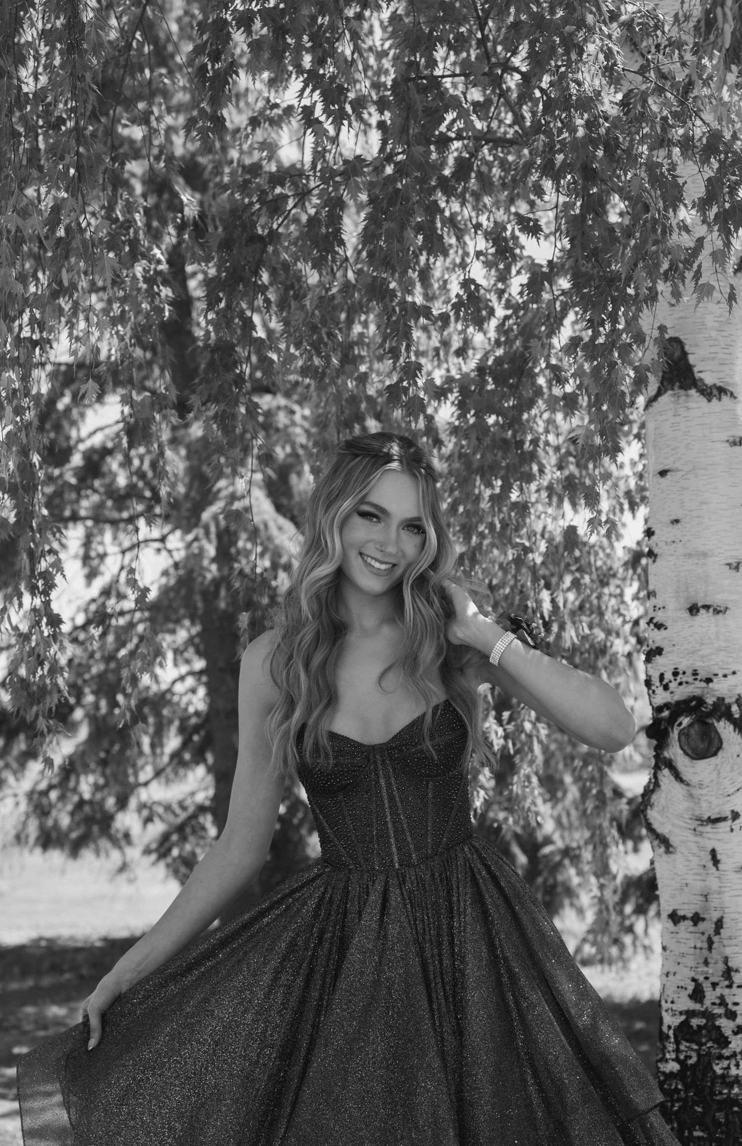 Black and white portrait of a woman in a dark dress posing under birch trees in a dreamy outdoor setting.