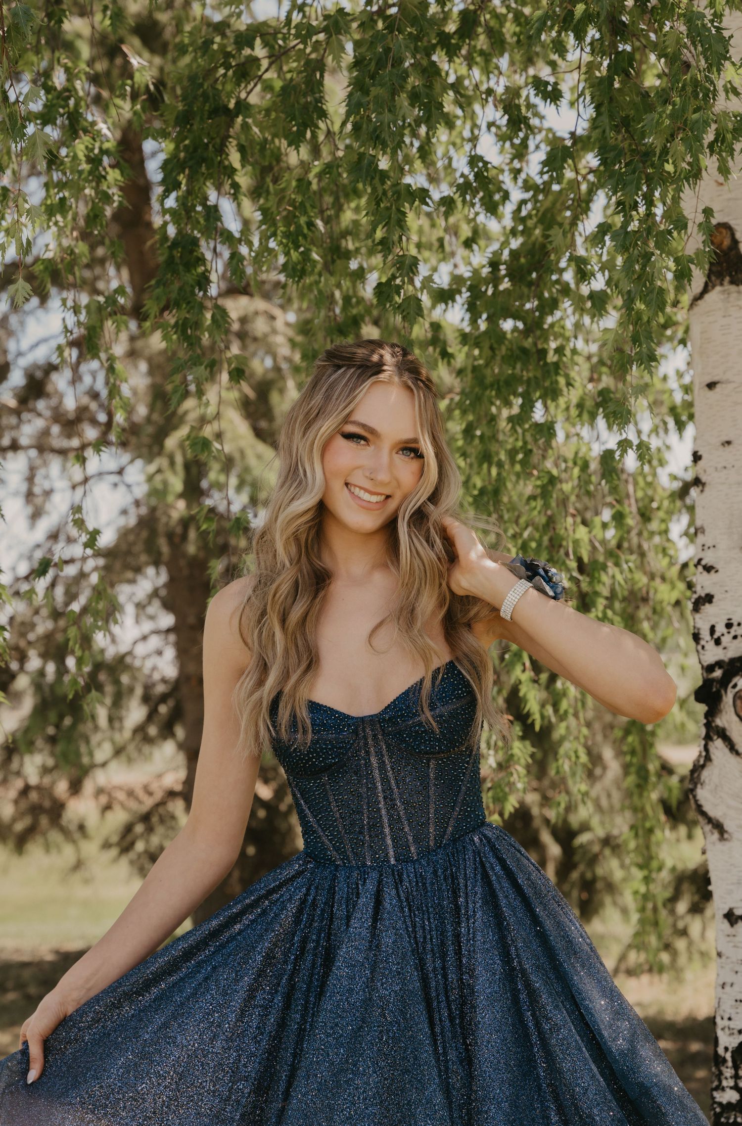 A person in a sparkly navy blue dress poses outdoors under hanging tree branches on a sunny day.