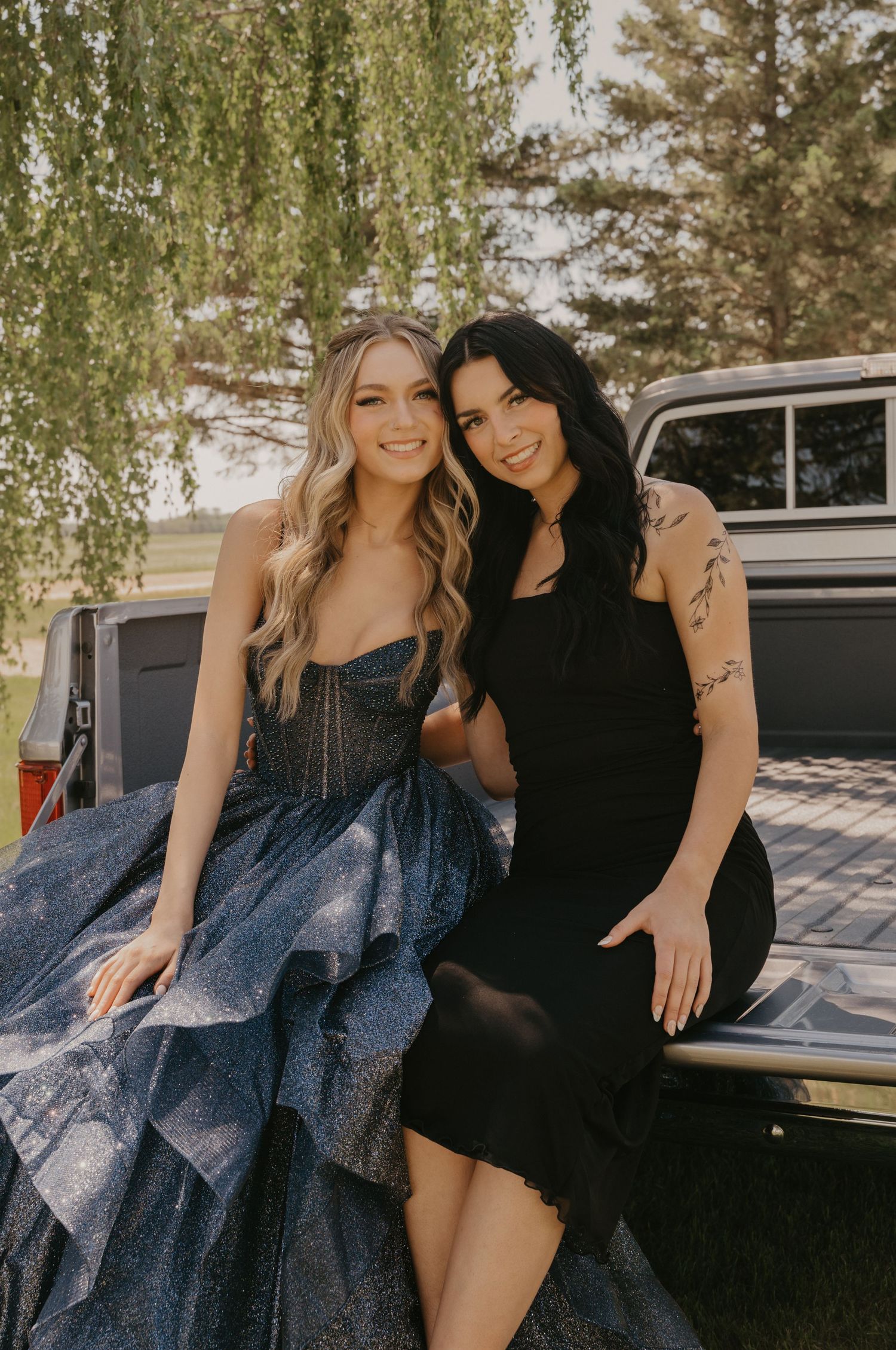 Two friends in formal attire pose together against a vintage car outdoors on a sunny day.