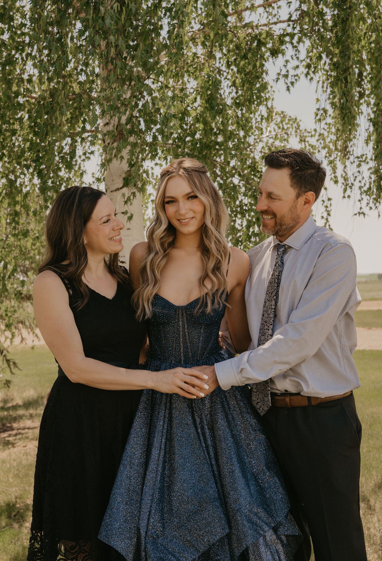 Three people stand together under a willow tree, with the center person wearing a navy blue formal gown.