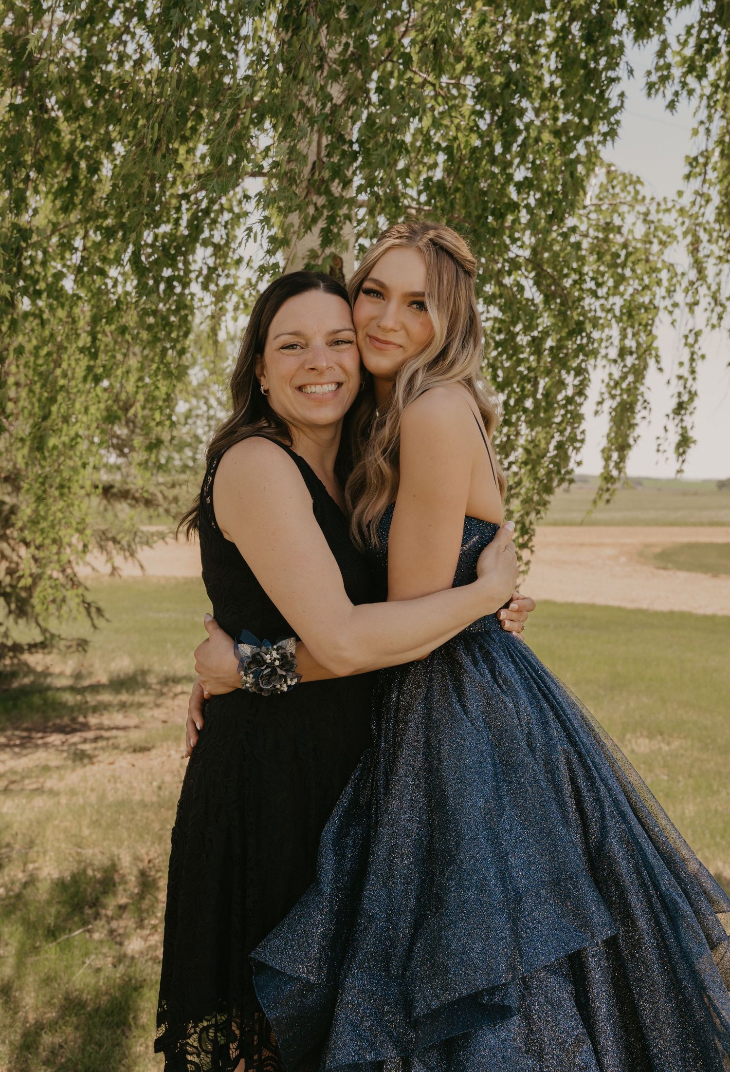 Two women in contrasting dresses embrace and smile under a willow tree in warm natural lighting.