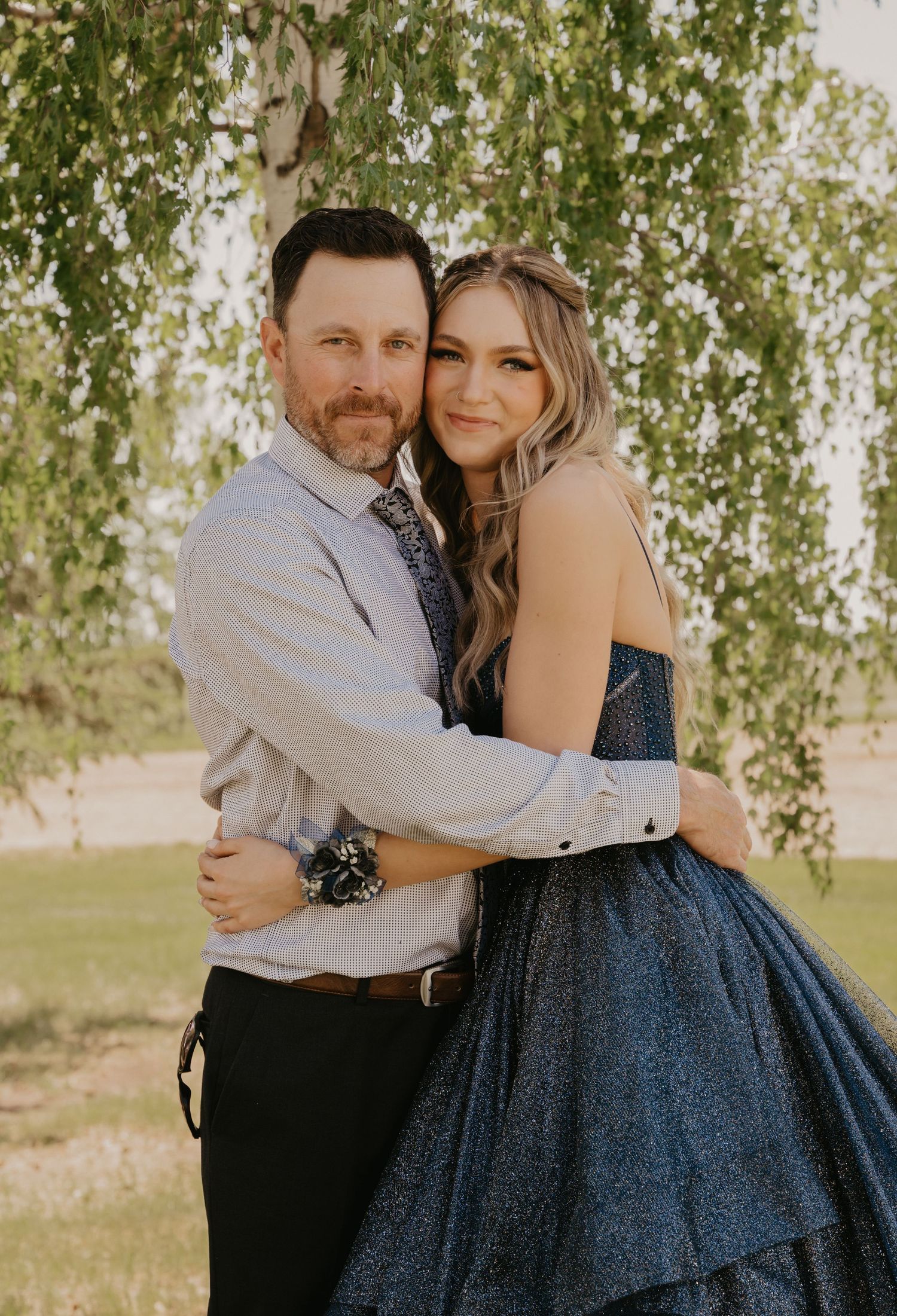 A romantic couple embraces under a willow tree, with the woman wearing a navy blue formal dress and the man in business attire.