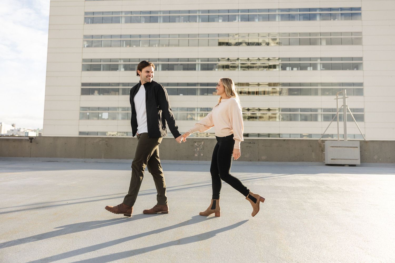 Stylish couple walking hand in hand on a rooftop parking lot with a modern building backdrop.