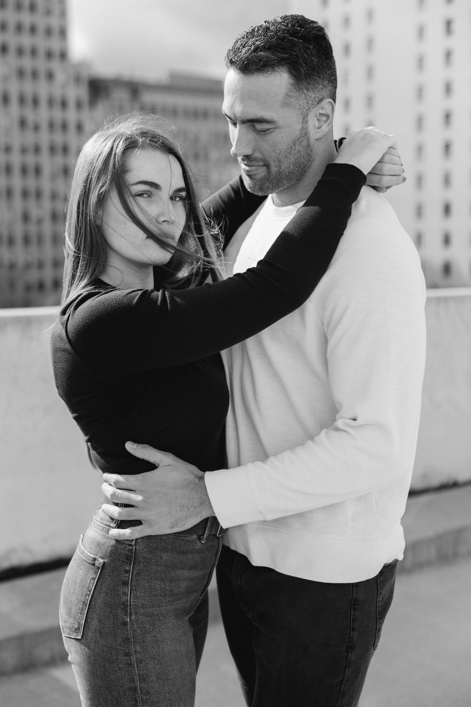 Black and white photo of a loving couple embracing on a rooftop with city buildings in the background.