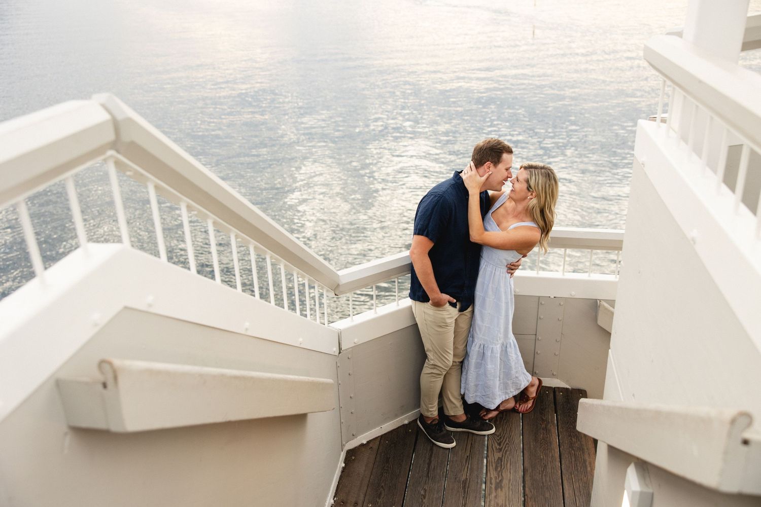 Couple sharing a romantic kiss on white waterfront stairs with sparkling ocean in the background.