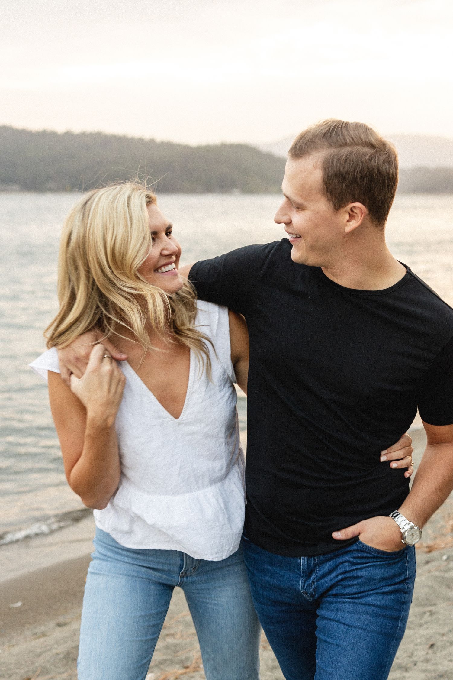 Happy couple laughing together on a sandy beach at sunset, woman in white top and jeans.